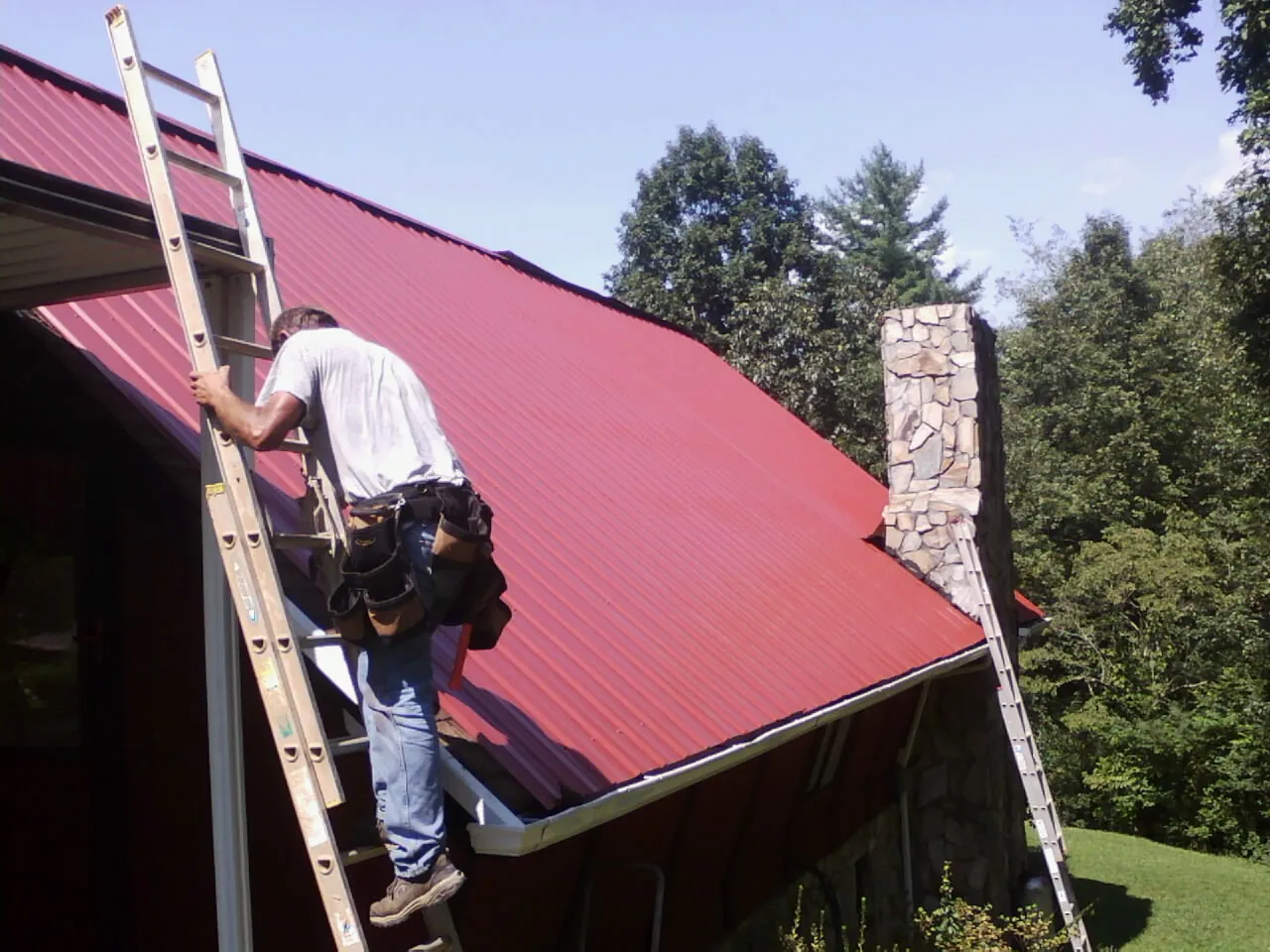 Roofer on a ladder beside a red metal roof and stone chimney — Soffit & Fascia Repair by Roofing Brothers in Nunnelly